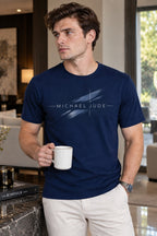 A man wearing a Michael Jude navy blue crewneck t-shirt with a white brushstroke graphic logo, holding a coffee mug.