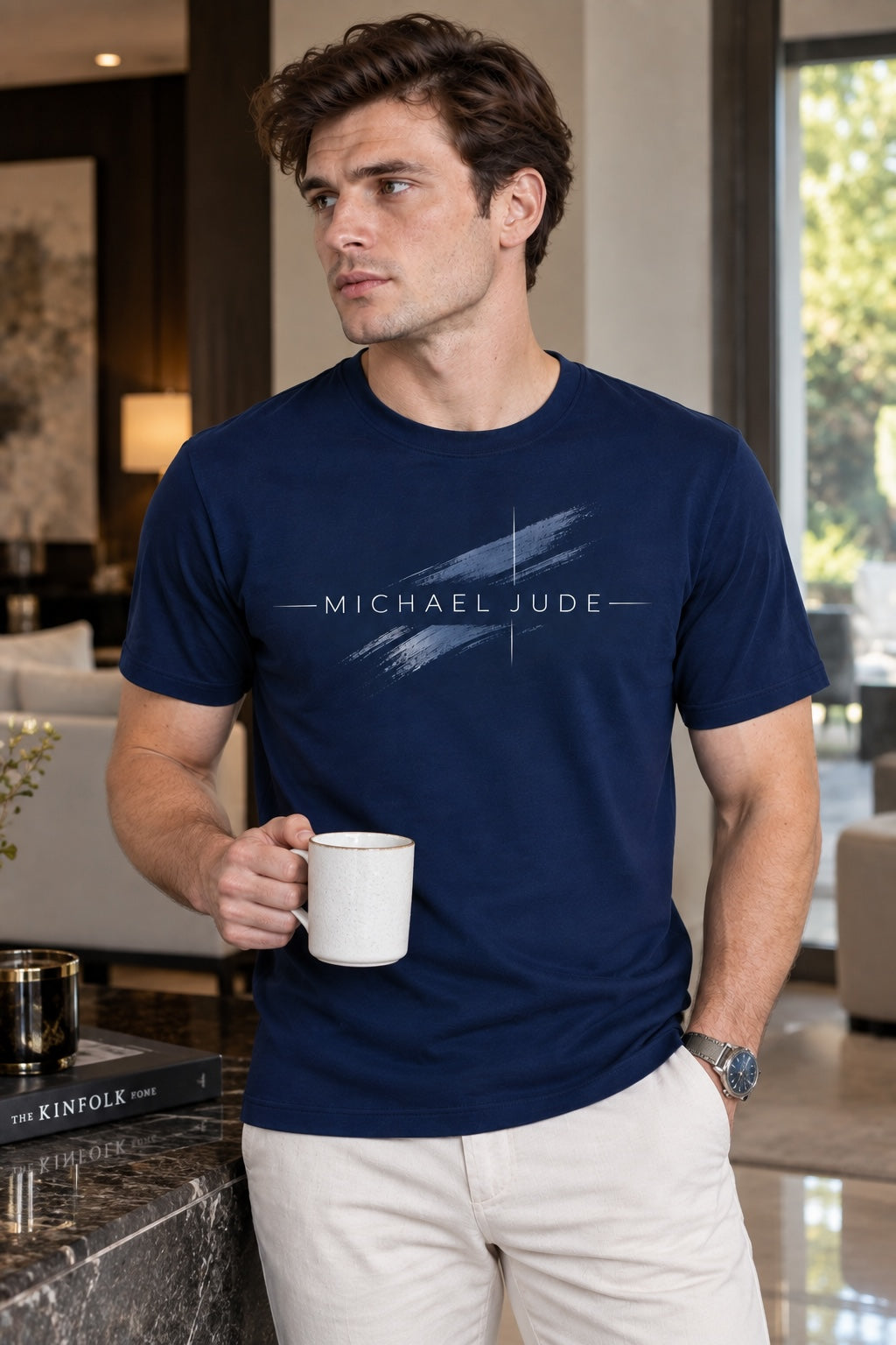 A man wearing a Michael Jude navy blue crewneck t-shirt with a white brushstroke graphic logo, holding a coffee mug.